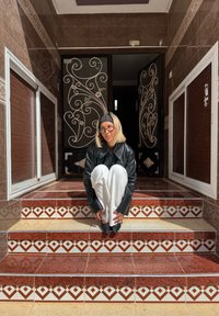 Persona con capelli biondi, occhiali, fascia nera per capelli, giacca di pelle e pantaloni bianchi accovacciata su gradini piastrellati con motivi davanti a porte ornate in metallo nero.