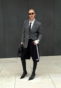 Grauer Blazer über einem weißen Hemd und schwarzer Krawatte, schwarze Sportshorts mit weißen Streifen, schwarze knielange Stiletto-Stiefel, schwarze Handtasche, Sonnenbrille.