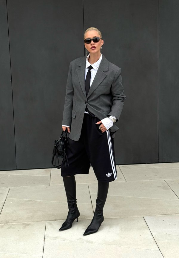 Šedý blazer přes bílou košili a černou kravatu, černé sportovní kraťasy s bílými pruhy, černé vysoké střevíce na jehlách po kolena, černá kabelka, sluneční brýle.
