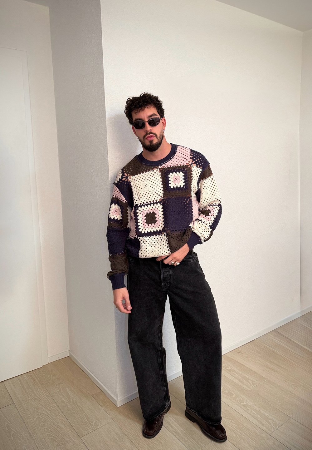 Suéter con patchwork en tonos crema, azul marino, rosa y marrón, combinado con jeans anchos negros y zapatos negros. Cabello rizado y gafas de sol.