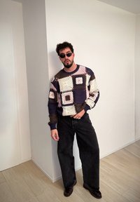 Suéter con patchwork en tonos crema, azul marino, rosa y marrón, combinado con jeans anchos negros y zapatos negros. Cabello rizado y gafas de sol.