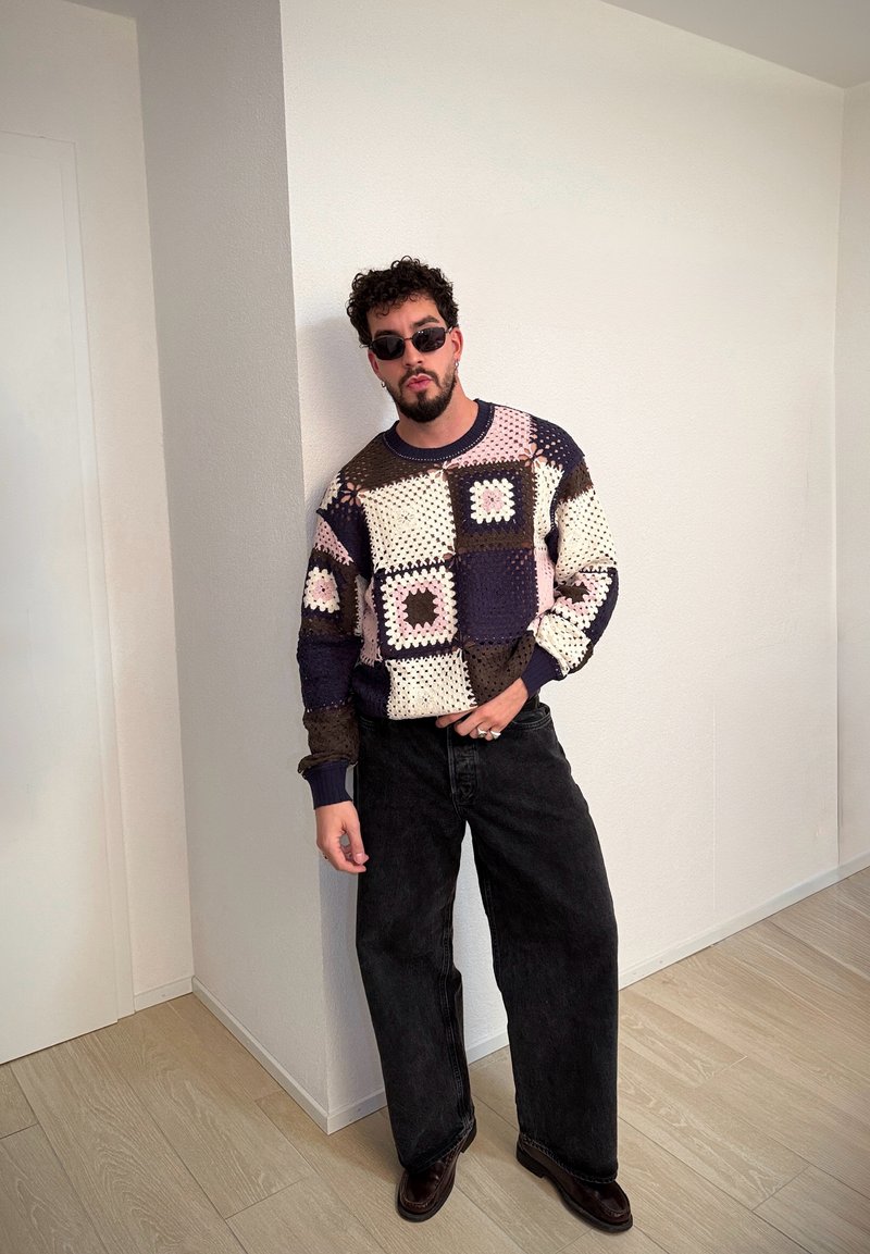 Pull-over avec patchwork dans des tons crème, marine, rose et marron, associé à un jean large noir et des chaussures noires. Cheveux bouclés et lunettes de soleil.