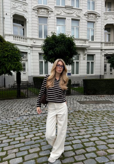 Top de rayas blanco y negro de manga larga, pantalones beige de pierna ancha y zapatillas grises. Bolsa marrón. Calle empedrada y fondo de vegetación.