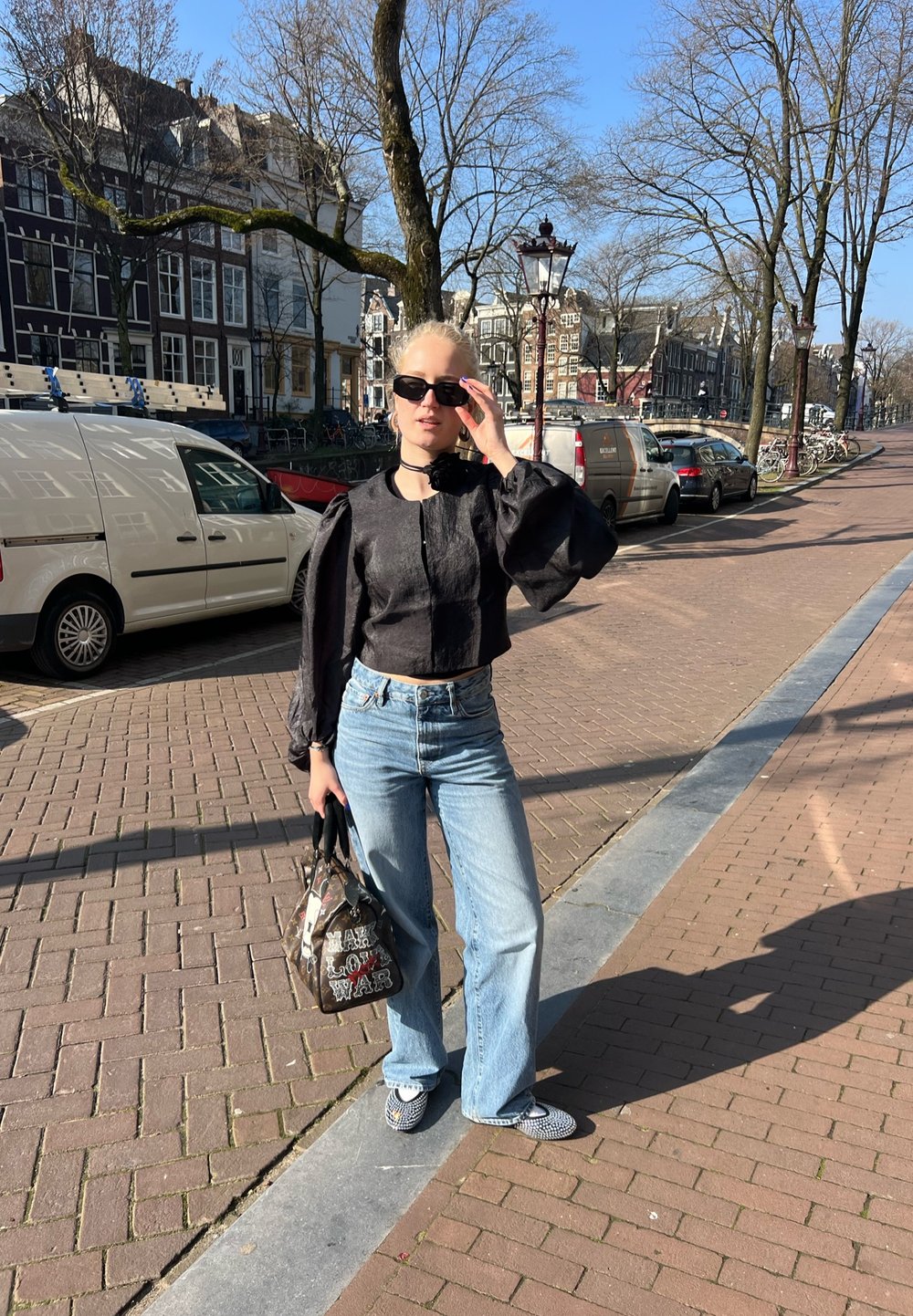 Črn skrajšan top s pufastimi rokavi in okroglim izrezom, v kombinaciji s svetlobluzimi širokimi hlačami. Dodatki vključujejo sončna očala in torbico z vzorcem.