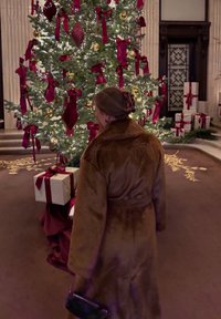 Brun pelsfrakke med stor krave, stående nær et juletræ pyntet med røde bånd og guldornamenter, omgivet af indpakkede gaver.
