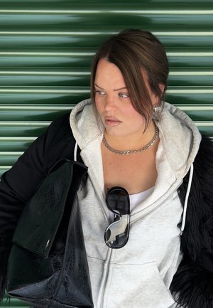 Mujer con sudadera con capucha gris y chaqueta negra, sosteniendo una gran bolsa negra, con gafas de sol colgando de la cremallera de la sudadera, frente a un fondo verde corrugado.