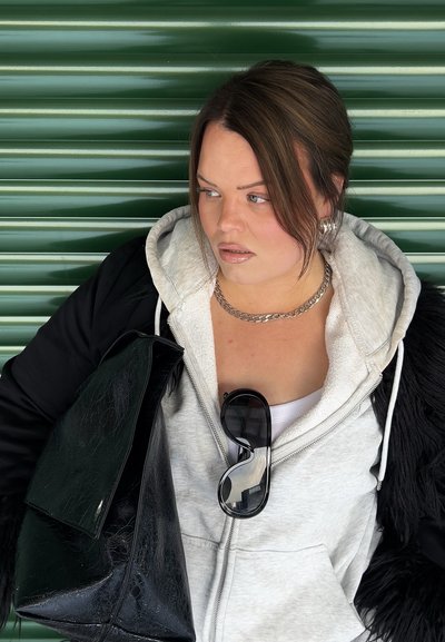 Mujer con sudadera con capucha gris y chaqueta negra, sosteniendo una gran bolsa negra, con gafas de sol colgando de la cremallera de la sudadera, frente a un fondo verde corrugado.