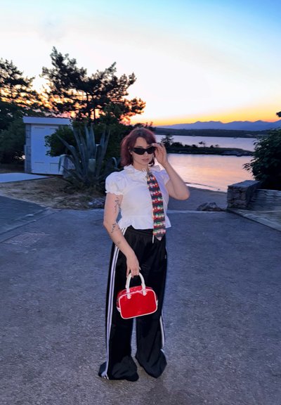 Blusa corta blanca con mangas abullonadas, corbata estampada, pantalones deportivos negros con rayas blancas y un pequeño bolso rojo. Fondo de atardecer.