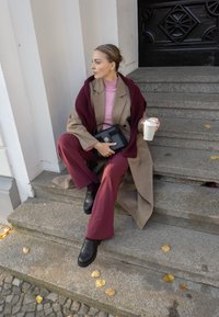 Casaco bege, suéter bordo, calças burgundy, sapatos de loafer pretos, segurando uma chávena de café branca, sentado em degraus de pedra com folhas de outono. Bolsa preta.