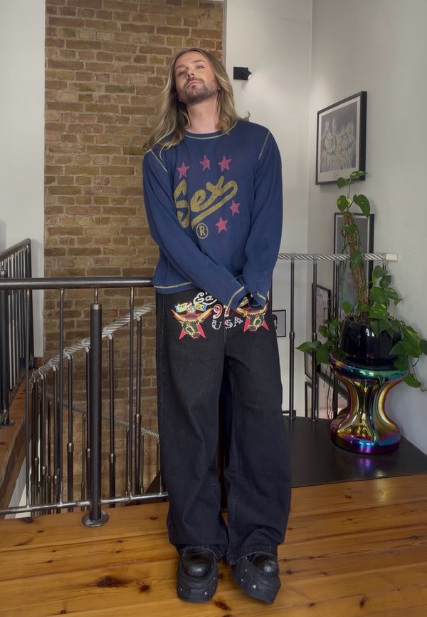 Personne aux cheveux longs portant un pull bleu et un pantalon noir décoré se tient à l'intérieur à côté d'un escalier et d'un pot de plante coloré sur un sol en bois.
