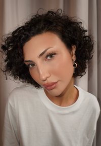 Žena s krátkymi kučeravými tmavými vlasmi a kruhovými náušnicami, oblečená v bielej košeli, prikláňa hlavu mierne k béžovej záclone v pozadí.