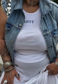 Hvitt ribbet tank-top med "LEVI'S" logo, parret med en lyseblå denimvest med sølvpaljetter på skuldrene. Hvit teksturert skjørt synlig.