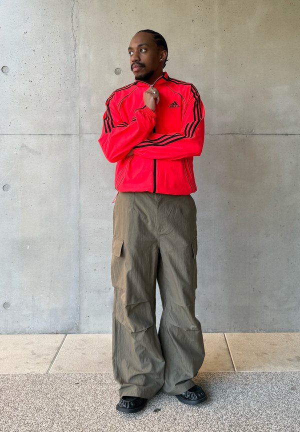 Mężczyzna z plecionymi włosami, ubrany w jaskrawoczerwoną kurtkę Adidas i szerokie oliwkowe spodnie typu cargo, stoi przy betonowej ścianie, patrząc na bok.