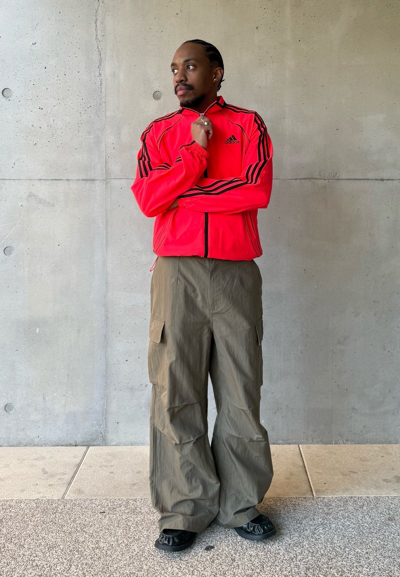 Άνδρας με πλεξούδες, φοράει ένα φωτεινό κόκκινο μπουφάν Adidas και φαρδιά ελιά cargo παντελόνια, στέκεται απέναντι σε έναν τοίχο από σκυρόδεμα, κοιτώντας προς τα πλάγια.