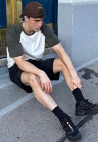 Schwarze Shorts, weißes und olivfarbenes T-Shirt, schwarze Socken und schwarze Sneaker mit bunten Akzenten. Der Hintergrund zeigt eine blaue Tür und graue Stufen.