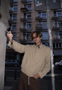 Bēša rāvējslēdzēja jaka ar augstu apkakli virs melnas krekliņa, brūnas bikses un saulesbrilles, turot kameru pie staba pilsētas vidē.