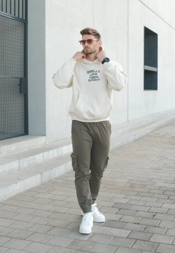 Homme portant des lunettes de soleil ajuste la capuche d’un sweat crème sur lequel est inscrit "Smells Like Teen Spirit" en marchant sur un trottoir pavé.