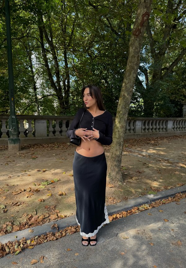 Femme portant un haut court noir et une longue jupe, debout sur un chemin feuilleux près d'un arbre et d'une balustrade en pierre dans un parc ombragé.