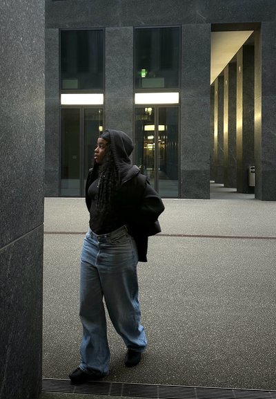 Sudadera negra, camiseta negra, jeans de pierna ancha de color azul claro y zapatos negros. Edificio de piedra gris, grandes ventanales de vidrio y pavimento texturizado.