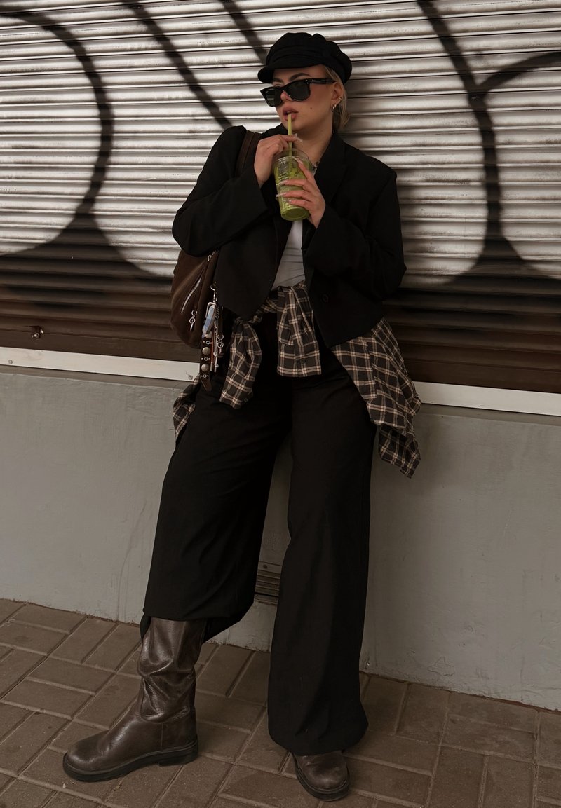 Eine Person in einem schwarzen, übergroßen Blazer, weißem Hemd und einem karierten Flanellhemd, das um die Taille gebunden ist. Sie hält ein grünes Getränk und trägt dunkle Sonnenbrillen.