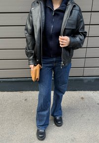 Veste en cuir noir à surface texturée, polaire marine zippée, jean large bleu, et chaussures épaisses noires. Tenant une pochette tissée marron.