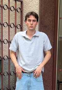 Camisa polo cinza com colarinho, combinada com jeans de ganga azul claro. O fundo apresenta uma parede estampada e um portão de metal.