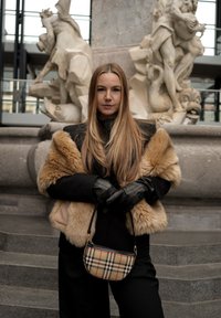 Γυναίκα με μακριά μαλλιά που φοράει μαύρο παλτό, γούνινο κασκόλ, δερμάτινα γάντια, κρατώντας τσάντα με καρό μοτίβο, στέκεται σε πέτρινα σκαλοπάτια μπροστά από ένα μεγάλο άγαλμα.