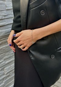 Pulseira de corrente prateada com elos em forma de clipe de papel e um charme floral, usada numa mão com unhas longas e azul-escuro, ao lado de um blaser de couro preto.