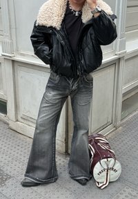 Black leather puffer jacket with a white shearling collar, teamed with flared grey jeans and black ankle boots. A red and white duffle bag is nearby.