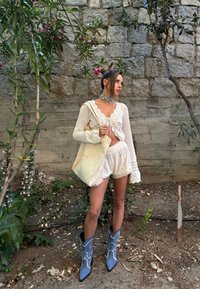 Pletena bela jopica in usklajene kratke hlače z čipkastim robom, v kombinaciji z modrimi kavbojskimi čevlji z belimi šivi in platneno torbo.