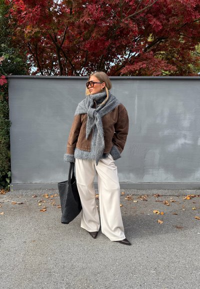 Chaqueta de ante marrón con bufanda gris y suave, pantalones anchos color crema y bolso tote negro tejido. Tacones marrones puntiagudos. Fondo de follaje otoñal.
