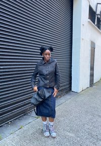 Giacca in denim nero con chiusura a bottoni, gonna in denim blu e scarpe da ginnastica grigie. Tiene una borsa nera. Sfondo con superficie scura e testurizzata.