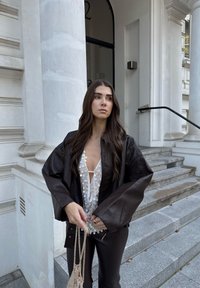 Ruskea nahkatakki, jossa on ylisuuret hihat, valkoisen blousen päällä, jossa on pyöreitä kuvioita. Malli pit�ää kädessään kudottua beigeä laukkua.