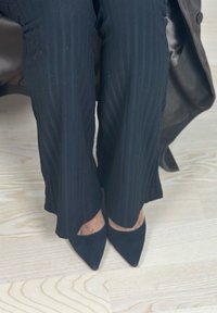 Personne portant un pantalon noir texturé et des talons pointus noirs debout sur un sol en bois clair avec une partie d'un manteau noir visible.