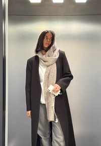 Kvinne i lang mørk frakk og beige skjerf, som holder en hvit smarttelefon med ørepropper, stående mot en enkel metallvegg.