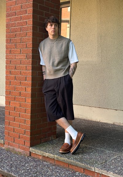 Mocasines marrones, calcetines blancos, pantalones cortos negros, camiseta sin mangas de punto gris claro sobre una camisa blanca. De pie junto a una columna de ladrillo contra una pared texturizada.