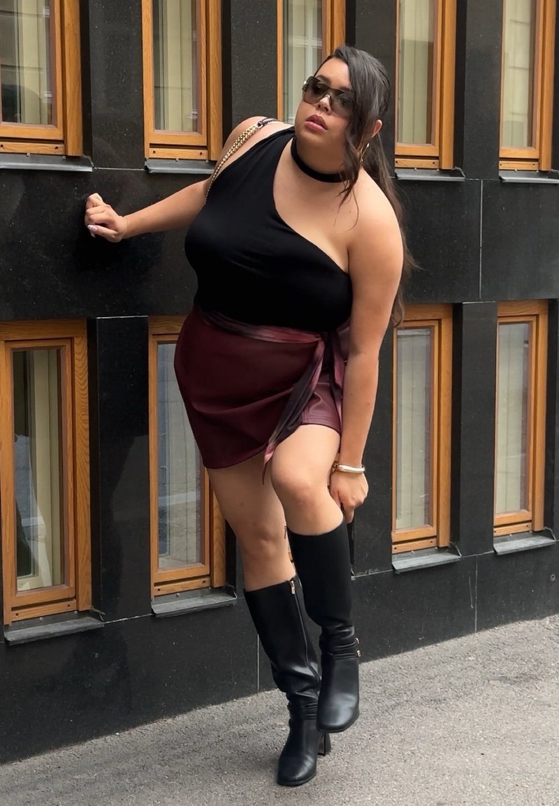 Femme portant des lunettes de soleil, un haut noir à une seule épaule, une jupe bordeaux et des bottes noires montantes, appuyée contre un mur noir avec des fenêtres à cadre en bois.