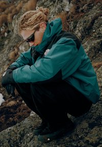 Τιρκουάζ και μαύρο μπουφάν με μόνωση και ψηλό γιακά, μαύρα γάντια και σκούρα υποδήματα, ποζαρισμένο μπροστά σε βραχώδες φόντο.