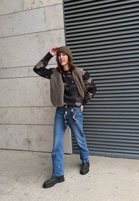 Lag-på-lag outfit med en blød beige vest over en camouflage-sweatshirt, blå jeans og mørke støvler, med en grøn strikket hue.