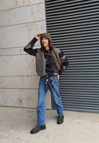 Conjunto en capas que incluye un chaleco beige peludo sobre una sudadera con capucha de camuflaje, vaqueros azules y botas oscuras, acompañado de un gorro de punto verde.