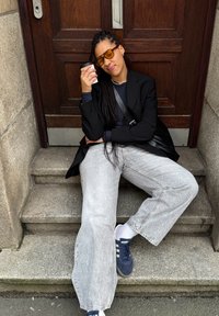 Blazer nero, top blu navy, jeans a gamba larga grigio chiaro, e sneakers blu navy. Indossa occhiali da sole gialli, tiene una tazza di caffè, seduto su gradini di pietra.