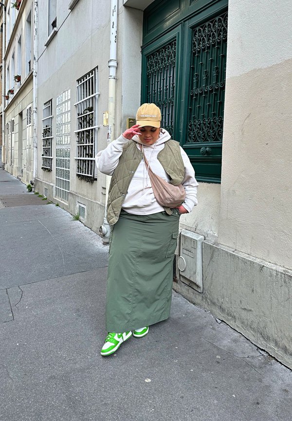 Lässiges Outfit mit einer beigen Mütze, grauem Hoodie, olivgrüner langer Rock und grünen Sneakers mit weißen Akzenten. Hintergrund: städtische Straße.