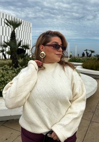 Κρέμα πλεκτή ζακέτα με διαμαντένιο σχέδιο, χαλαρή εφαρμογή, ψηλός γιακάς, φορεμένη με σκούρα γυαλιά ηλίου και χρυσά σκουλαρίκια, standing outdoors.