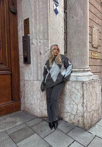 Femme aux longs cheveux blonds portant un grand pull à losanges et un jean foncé, appuyée contre un pilier d'angle en marbre à l'extérieur d'un bâtiment avec une porte en bois.