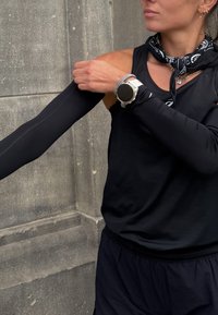 Débardeur de sport noir avec un col rond, associé à des manches longues noires. Une montre argentée et un bandana noir sont visibles.