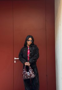 Veste courte en velours noir, pull à losanges rouge et gris, pantalon noir, et sac à main bordeaux avec des clous blancs, devant une porte rouge.
