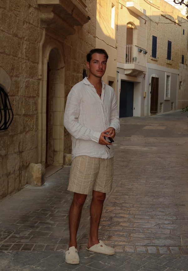 Helles Leinenhemd mit Knöpfen, gestreifte beigen Shorts und helle Clogs. Steht auf einer Steinstraße mit Gebäuden im Hintergrund.