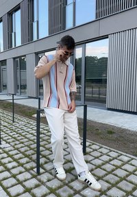 Kortærmet polo-shirt i beige med blå og røde striber, kombineret med hvide bukser og hvid sneakers med sorte striber. Udendørs scene.