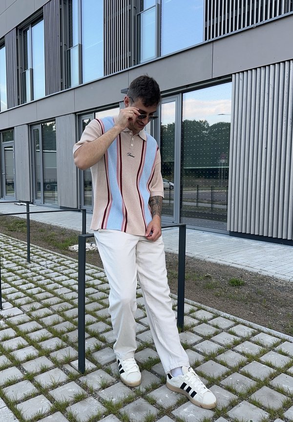 Korte mouwen polo in beige met blauwe en rode strepen, gecombineerd met witte broek en zwart gestreepte witte sneakers. Buitensetting.