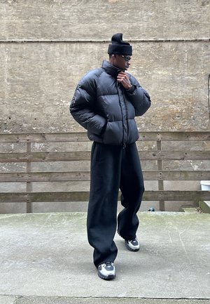 Schwarze Pufferjacke mit hohem Kragen, kombiniert mit lockeren schwarzen Hosen. Das Outfit umfasst metallisch-silberne Turnschuhe und eine schwarze Strickmütze.